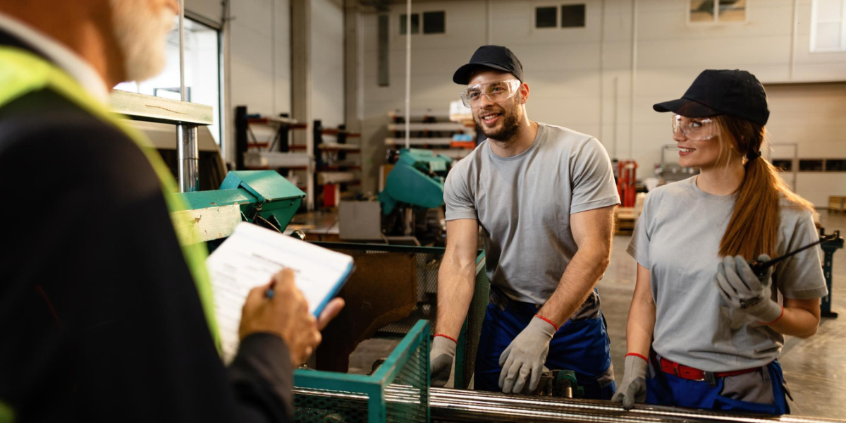 Two happy workers working at the machine and communicating with senior businessman who is overseeing them in a manufacturing building.