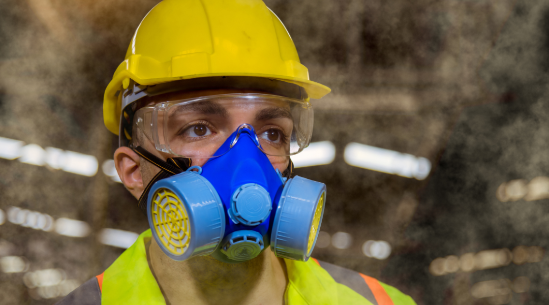 Man in a yellow hard hat with a yellow and organge safety vest on with clear safety goggles and a blue respirator mask on face
