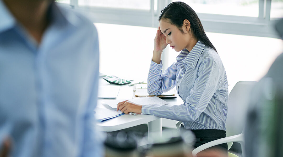 woman staring at documents at desk look stressed, people blurred in front of her
