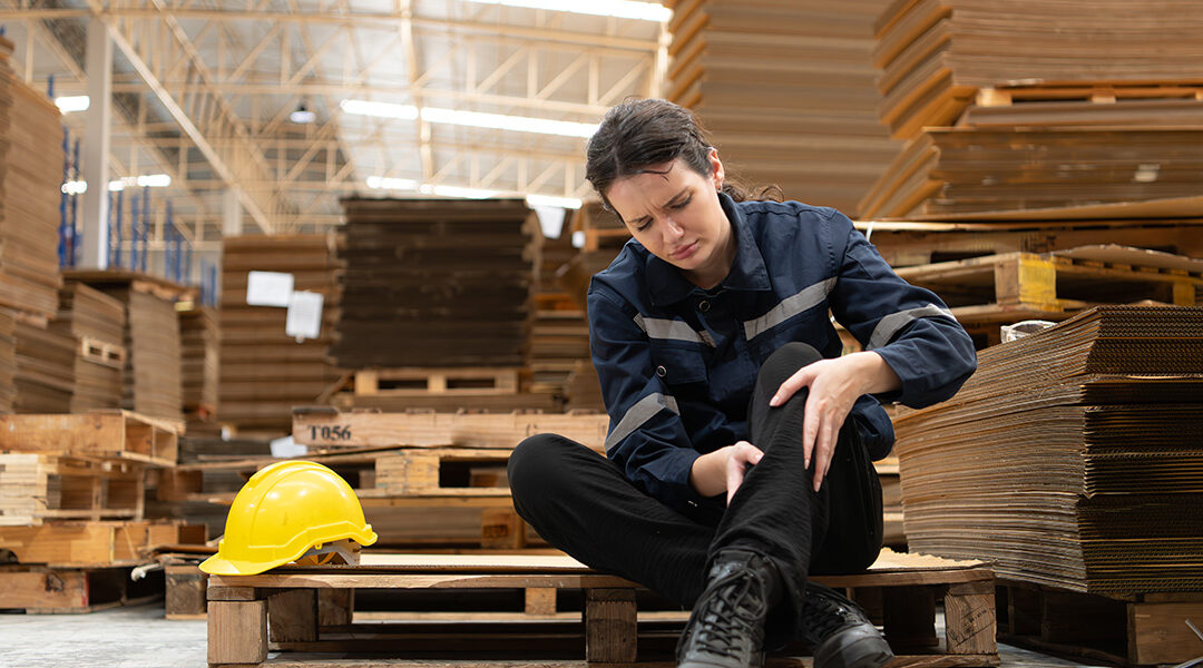 Warehouse worker sits on a pallet in a large facility, clutching her knee in pain, with a yellow hard hat beside her.