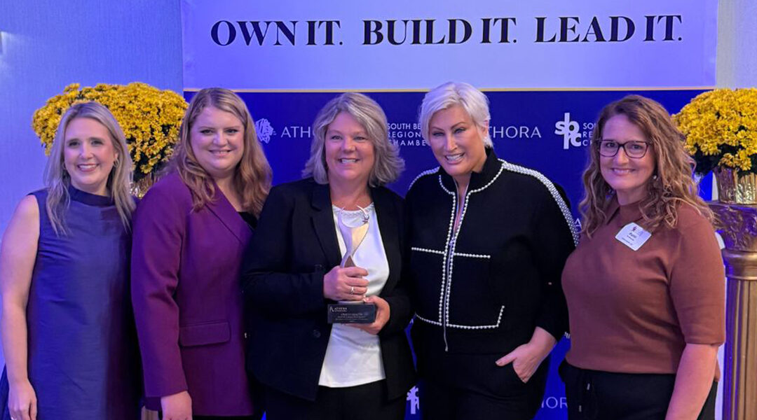 Five women in business attire pose at an awards event, one holding a trophy, with a blue banner reading 'OWN IT. BUILD IT. LEAD IT.' and yellow flower arrangements behind them.