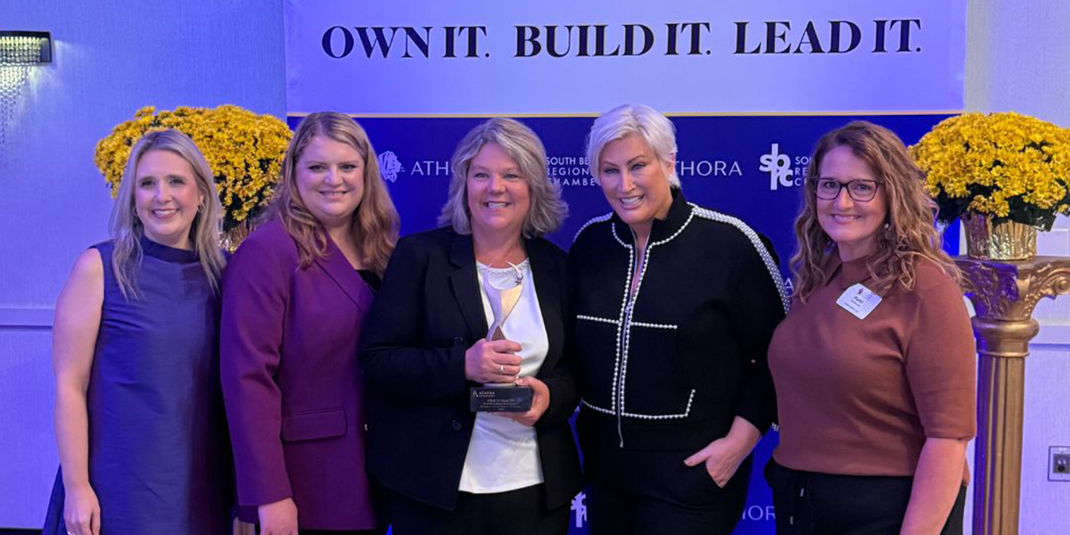 Five women in business attire pose at an awards event, one holding a trophy, with a blue banner reading 'OWN IT. BUILD IT. LEAD IT.' and yellow flower arrangements behind them.